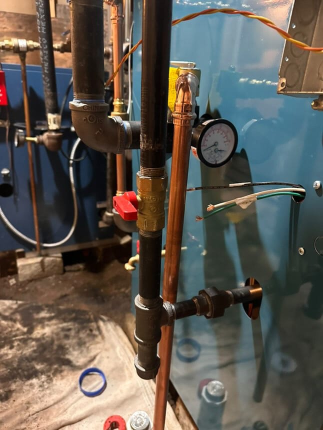 Gas boiler system with black iron and copper piping, pressure gauge, and shut-off valve in a mechanical room.