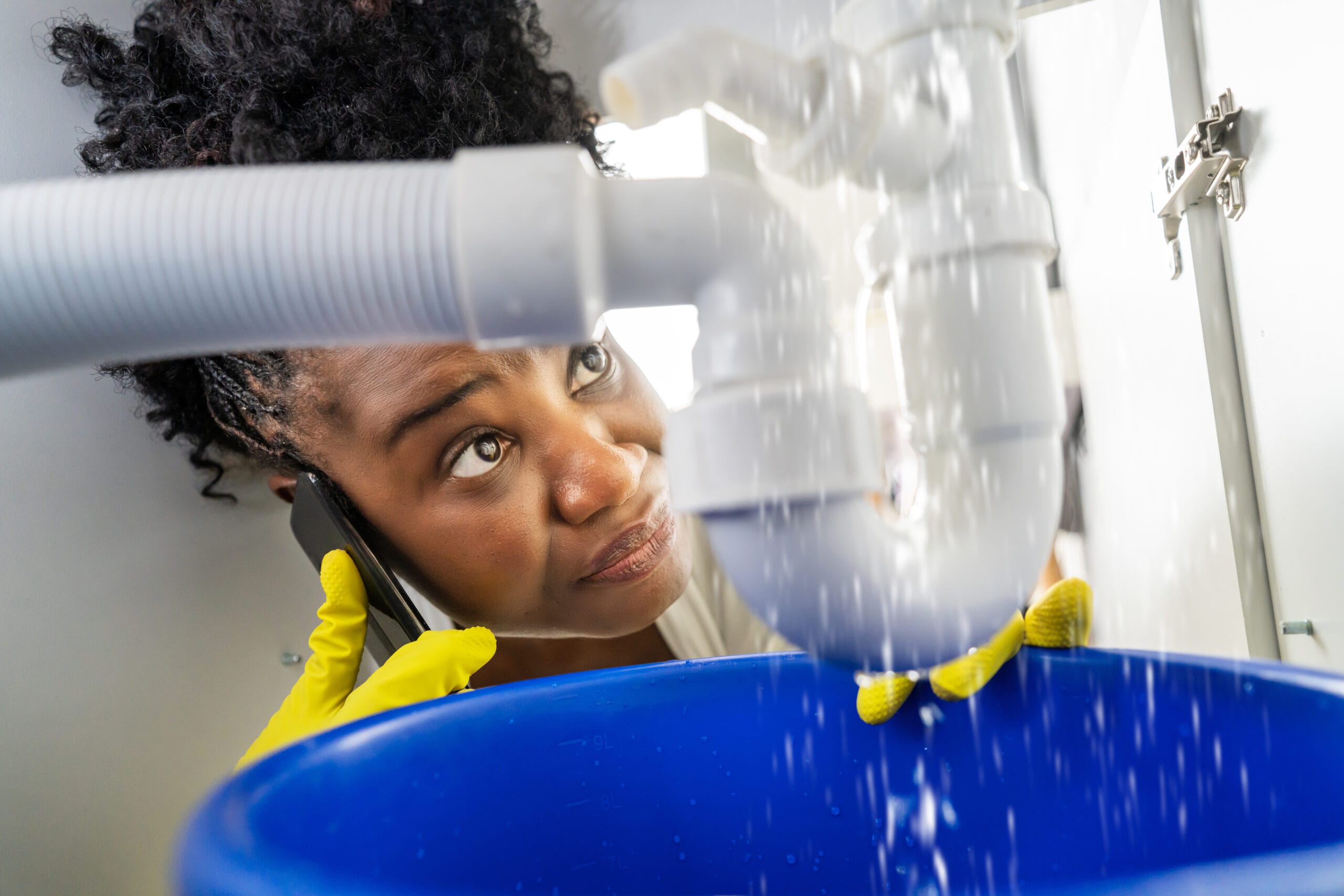 Woman wearing yellow gloves catching water from a leaking sink pipe while calling a plumber for emergency plumbing repair.
